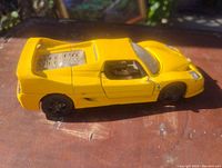 Side view photo showing yellow Ferrari toy car with visible rear engine details and black wheels on wooden surface.