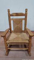 Front view of a child's wooden rocking chair with woven seat and backrest, showing spindle detail on posts and finial tops.