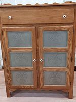 Front view showing double wood doors with six punched tin panel decorations and one top drawer with white ceramic knobs.