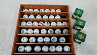 Top-down view of a wooden wall mount holder containing 36 golf balls arranged in five rows, each ball featuring different logos or motifs. Three golf ball boxes are positioned to the right of the holder.