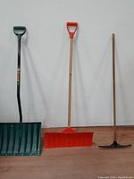 Full view of two plastic snow shovels and one metal rake leaning against the wall showing blades, handles and overall condition.