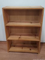 Front view of 3-shelf natural unfinished solid wood bookcase on wooden floor against white wall.