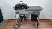 Full front view of the Weber Performer grill showing side table, charcoal bin, temperature gauge on lid, storage rack with firewood, and wheels.