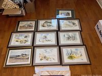 Photo shows 10 framed watercolour landscape prints arranged neatly on a hardwood floor, capturing the lot's contents and condition. Frames are brown with white mats.