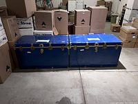 Two blue metal trunks side by side on concrete floor among cardboard boxes.