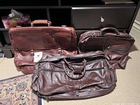 Three leather bags are shown: two dark brown and one lighter brown Hidesign briefcase-style bag with strap buckles, all placed on carpet in front of dark furniture.