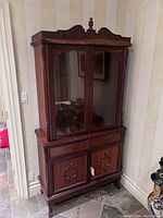Side angle photo showing the full height carved wooden hutch cabinet with glass doors and closed lower cabinet doors.