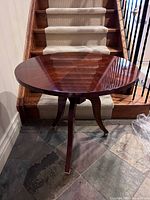 Round mahogany wood accent table with tripod base positioned in front of carpeted stairs, showing the tabletop and legs clearly.