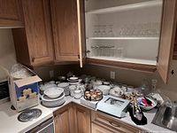 Overview of assorted kitchen items placed on countertop and inside cabinet, showing dinnerware, glassware and kitchen tools.