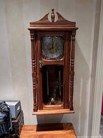 Front view of the wall mounted pendulum clock showing wood frame, clock face with Arabic numerals, and visible pendulum inside a glass panel.