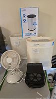 Two Honeywell air purifiers, small portable fan, Heat Machine Plus heater, and replacement HEPA filter in box on table.