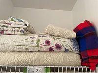 Stack of queen-sized bedspreads and shams on a wire shelf with Edmond acrylic plaid throw blanket folded and placed beside.