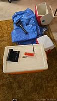 Photo showing the orange Coleman cooler with a white lid, blue tarps, a red and white cooler, black flashlight, black sunglasses, and a silver travel mug on a carpet.