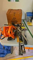 Full view of carpentry tools arranged on table showing circular saw, hammer, level, tri-square, wooden toolbox and crate of nails