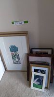Photo showing multiple framed art pieces stacked against a wall, varying sizes and frame types