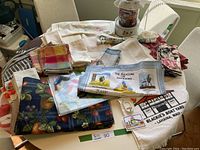 Lot of assorted kitchen linens including placemats, napkins, tea towels, aprons and tablecloths piled on a table.