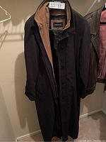 Black London Fog rain coat with brown interior lining shown buttoned and hanging on a hanger in closet.