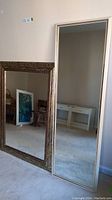Photo showing both mirrors standing near a wall, reflecting parts of the room. The floor-length mirror has a narrow light wood frame.