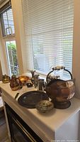 Wide angle showing the whole collection of copper, brass, and pewter decor pieces placed on a white surface near a window.