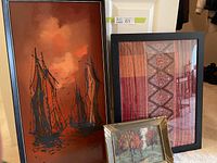 Full view of the three items: a tall framed painting of ships, a smaller fall landscape painting in a gold leaf frame, and a framed scarf with warm-toned textile patterns