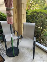 Two grey metal framed patio chairs with fabric seating seen on a concrete porch with a red patio umbrella and cast iron stand close by.