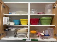 Two shelves inside a kitchen cabinet filled with colorful plastic mixing bowls, clear containers, a plastic sieve, and small kitchen accessories.