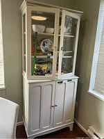 Front side view of the cream-colored step back cupboard showing the glass upper cabinet with shelves containing dishware and the closed lower cabinet doors.
