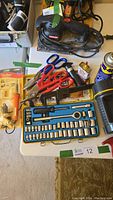Image of the lot showing socket set, shears, pipe wrench, soldering tool, oils, and other hand tools on a table.
