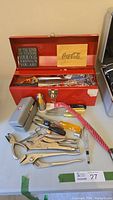 Photo of open red steel toolbox on table showing assortment of hand tools including pliers, wrenches, screwdrivers, saws, and files, along with decorative dog and Coca-Cola plaques inside lid.