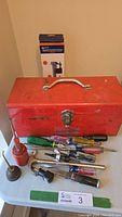 Front view of red steel toolbox and hand tools including oil can and various screwdrivers on table surface.