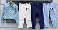 Four Calvin Klein women's clothing items hanging on rack: light wash denim jacket on far left, followed by two pairs of cropped pants in cream and navy blue, and periwinkle blue pants on far right.