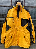 Front view of Helly Henson yellow and black ski jacket showing zippered and snap button pockets, front closure, and elastic waist detail.