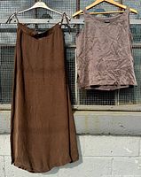 Photo showing the full view of the chocolate brown silky maxi skirt and taupe silk tank top hanging on wooden hangers against a wire mesh screen and concrete wall.
