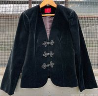 Full front view of black velvet women's blazer with three sets of frog fasteners, hanging on a wooden hanger outdoors.