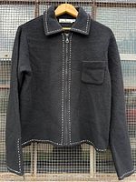 Front view of black André Christian wool jacket with cream blanket stitch detail showing collar, zipper, pocket, cuffs, and hem.