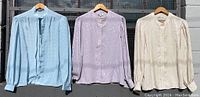 Front view of three vintage collared-less button-down women's blouses in light blue, lavender, and light peach colors hung on wooden hangers against a wall.