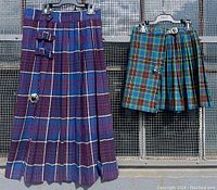 Two vintage women's plaid kilt skirts hanging on hangers, one short with green, blue, red plaid and a leather belt; one long blue and purple plaid