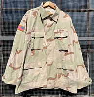 Full front view of vintage 1995 U.S. Army desert camouflage combat jacket hanging to show front pockets, collar, and sleeve patches under natural lighting.