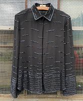 Front view of black sheer long sleeve top with bugle beading, hanging on wooden hanger against wire mesh background.