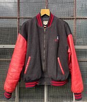 Front view of the varsity jacket hanging on hanger showing black wool body with red leather sleeves and striped ribbing on collar, cuffs, and hem.