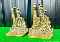 Pair of antique brass bookends depicting two lighthouses on rocky cliffs captured from the front and side angles, showing detailed casting and patina.