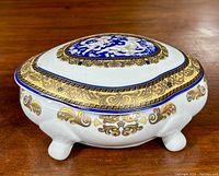 Porcelain trinket box showing top, side, and feet. Blue medallion with cherub relief on lid, gold and white ornate decoration on body.