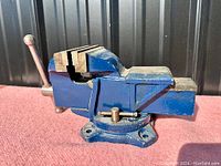 Front and side view of blue metal bench vise with handle and serrated jaws on a pink surface, with black background.