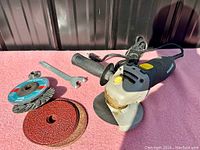 Mastercraft corded angle grinder with handle, replacement discs, wire brush disc, and wrench on pink fabric background