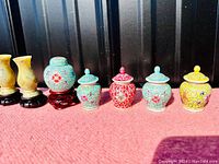 Photo of miniature porcelain ginger jars and carved soapstone vases on a pink surface with black background.