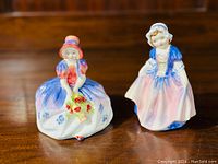 Two Royal Doulton figurines, "Monica" holding flowers and "Dinky Do" lifting her dress, displayed on wooden surface.