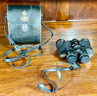 Black Tasco Model No. 210 7x35 binoculars with neck strap alongside a black carrying case with Tasco emblem and metal clasp, set on wooden surface.