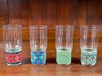 Four vintage MCM clear glass tumblers with colorful removable fabric bases in different patterns, displayed side by side on a wooden surface