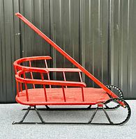 Side view of vintage red wooden sleigh showing curved wooden seat and spindled backrest with black metal runners.