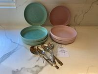 Pink and green Anthropologie plates stacked in two piles with four metal serving spoons laid in front, displayed on a marble countertop.